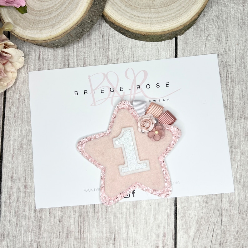 May include: Pink glitter star with a white felt number one and pink and white ribbon embellishments. The star is on a white card with the text "BRIEGE & ROSE WEAR" printed on it.