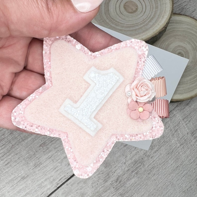 May include: Pink felt star with a white glitter number one and pink flower embellishments.