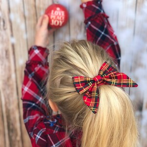 May include: A young girl with blonde hair wearing a red and black plaid shirt and a red and green plaid bow in her hair. She is holding a red Christmas ornament that says "Love Peace Joy".
