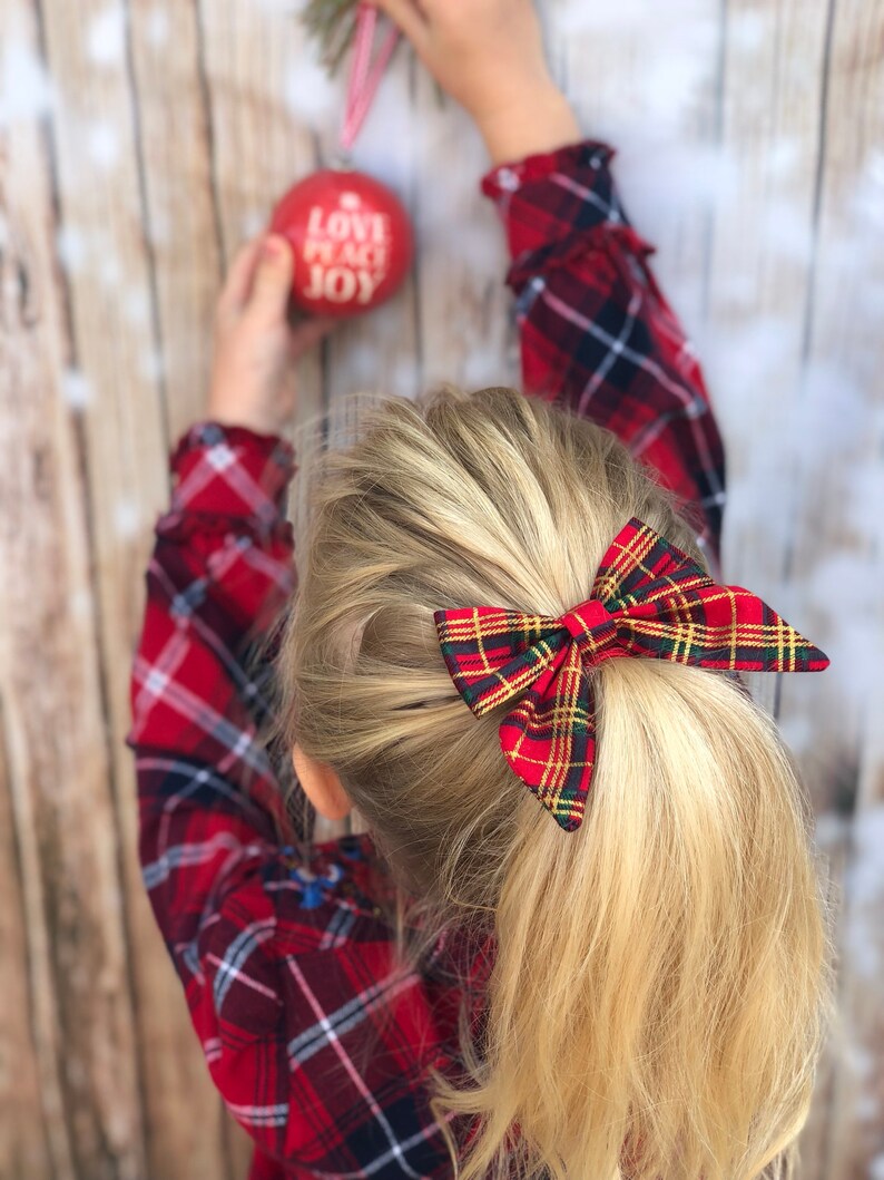 May include: A young girl with blonde hair wearing a red and black plaid shirt and a red and green plaid bow in her hair. She is holding a red Christmas ornament that says "Love Peace Joy".