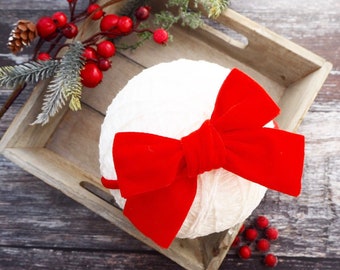 May include: A red velvet bow headband on a white background. The headband is made of a soft, plush velvet and has a large, oversized bow. The bow is tied in a knot at the center and the ends of the bow are left long and flowing. The headband is perfect for adding a touch of festive cheer to any outfit.