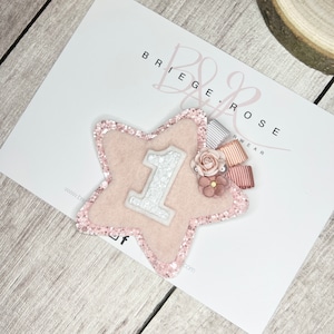 May include: Pink glitter star with a white felt number one, pink and white ribbon, and pink flowers.  The star is on a white background with the text "Briege & Rose Wear" in black.