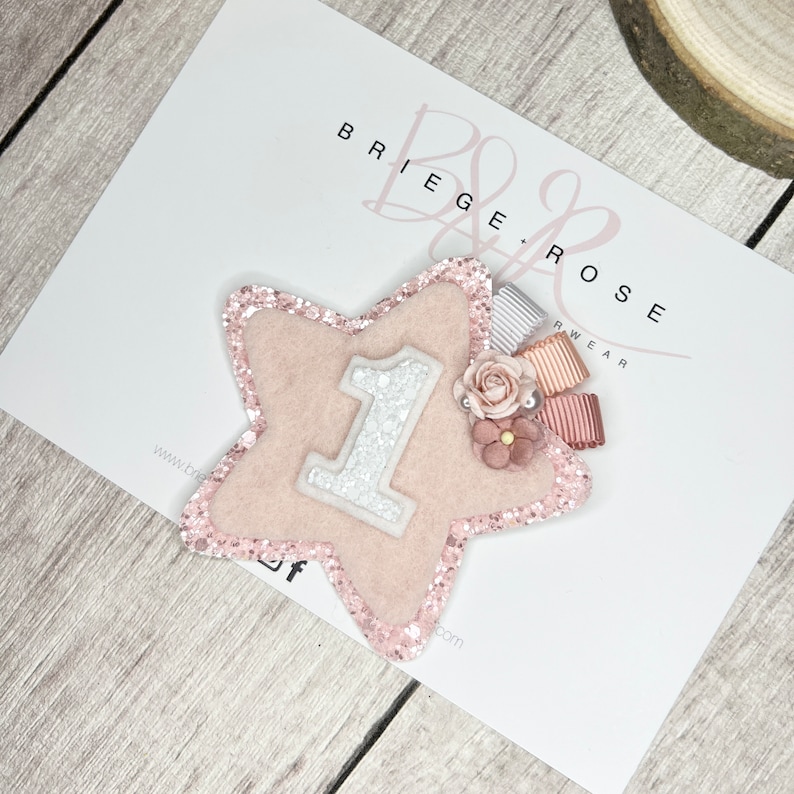 May include: Pink glitter star with a white felt number one, pink and white ribbon, and pink flowers.  The star is on a white background with the text "Briege & Rose Wear" in black.