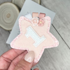 May include: Pink felt star with a white felt number one, decorated with pink and peach satin ribbon and pink and white felt flowers.