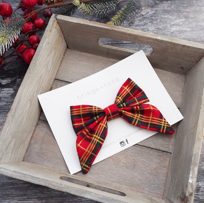 May include: Red and green plaid bow with gold accents. The bow is displayed in a wooden tray with the text "briege+rose" on a white card.