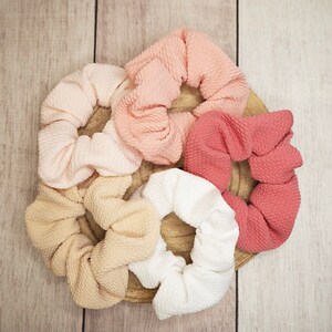 May include: Five fabric scrunchies in various shades of pink, peach, and beige. The scrunchies are made of a soft, textured fabric and are ideal for holding hair in place.