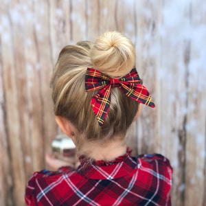 May include: A young girl with blonde hair in a bun, wearing a red and black plaid shirt, with a red and green plaid bow tied in her hair.