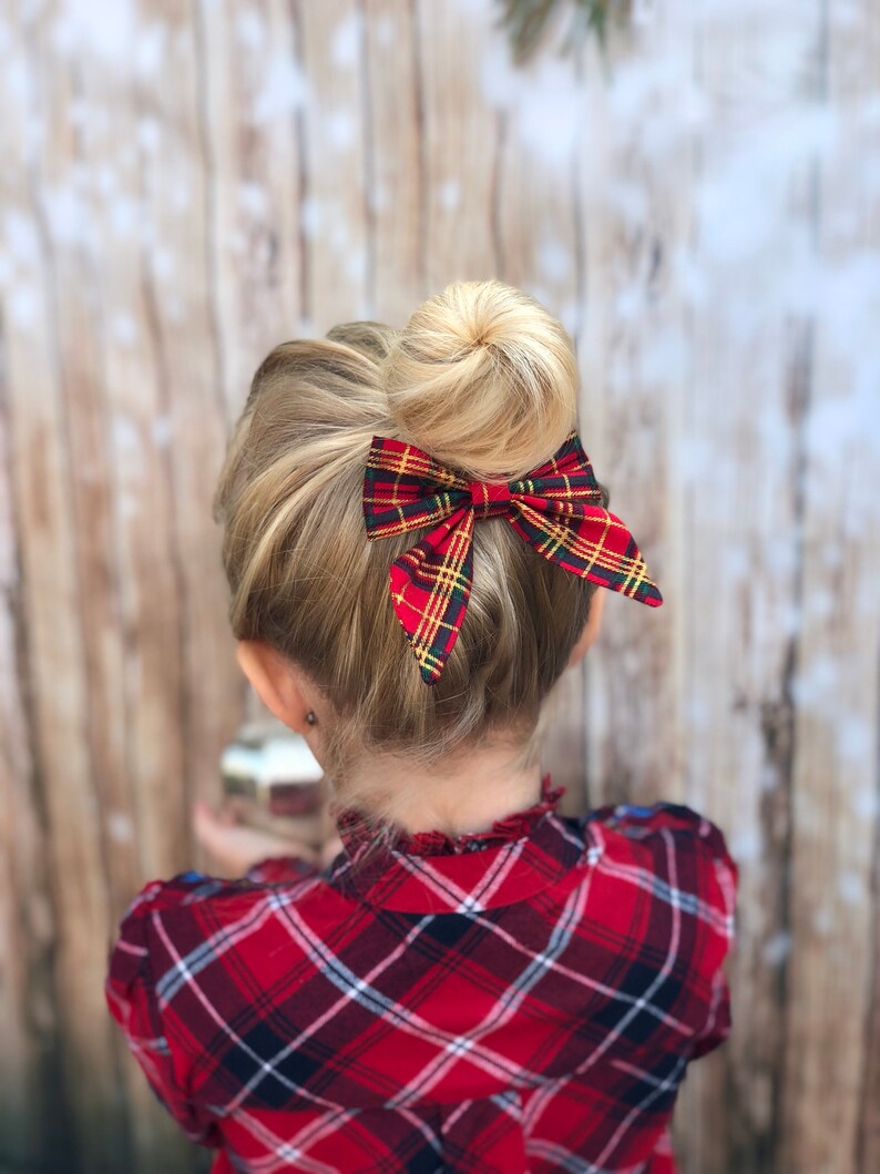 May include: A young girl with blonde hair in a bun, wearing a red and black plaid shirt, with a red and green plaid bow tied in her hair.