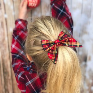May include: A red and green plaid hair bow with gold accents. The bow is tied in a ponytail.