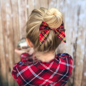 May include: A young girl with blonde hair wearing a red and black plaid shirt with a red and green plaid bow in her hair.