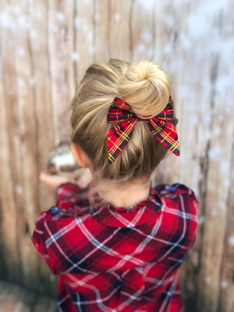 May include: A young girl with blonde hair wearing a red and black plaid shirt with a red and green plaid bow in her hair.
