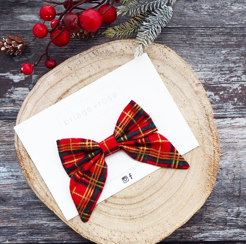 May include: A red and gold plaid bow with a white background. The bow is on a wooden surface with the text "briege+rose" on the white background.