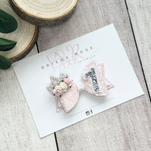 May include: Pink glitter bow with a silver glitter number one and a small white crown with pink flowers. The bow is on a white card with the text "Briege & Rose Hairwear" and a website address.