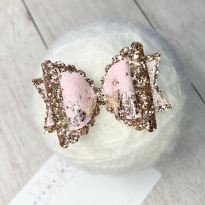 May include: A pink and gold glitter hair bow with a scalloped edge.