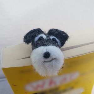 May include: A black and white felt miniature schnauzer dog bookmark. The dog is peeking over the top of a yellow book.