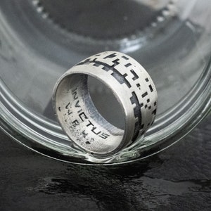 May include: A silver ring with a geometric pattern on the band. The ring is inscribed with the word "INVICTUS" on the inside of the band.