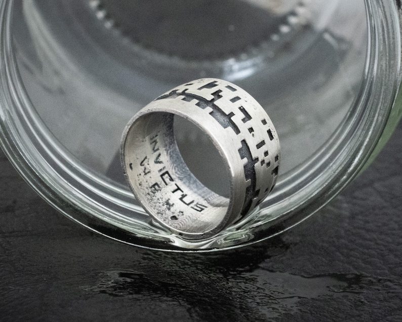 May include: A silver ring with a geometric pattern on the band. The ring is inscribed with the word "INVICTUS" on the inside of the band.