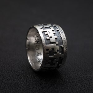 May include: A silver ring with a geometric pattern and inscription. The ring is on a black background.
