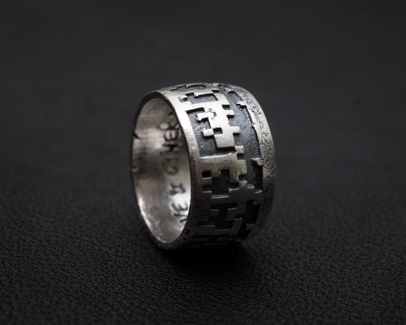 May include: A silver ring with a geometric pattern and inscription. The ring is on a black background.