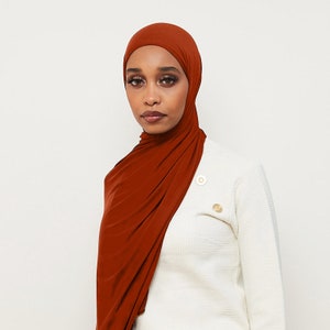 May include: A rust-colored jersey knit hijab with a simple, draped style. The hijab is worn by a person with dark skin and brown eyes. The person is wearing a white sweater.