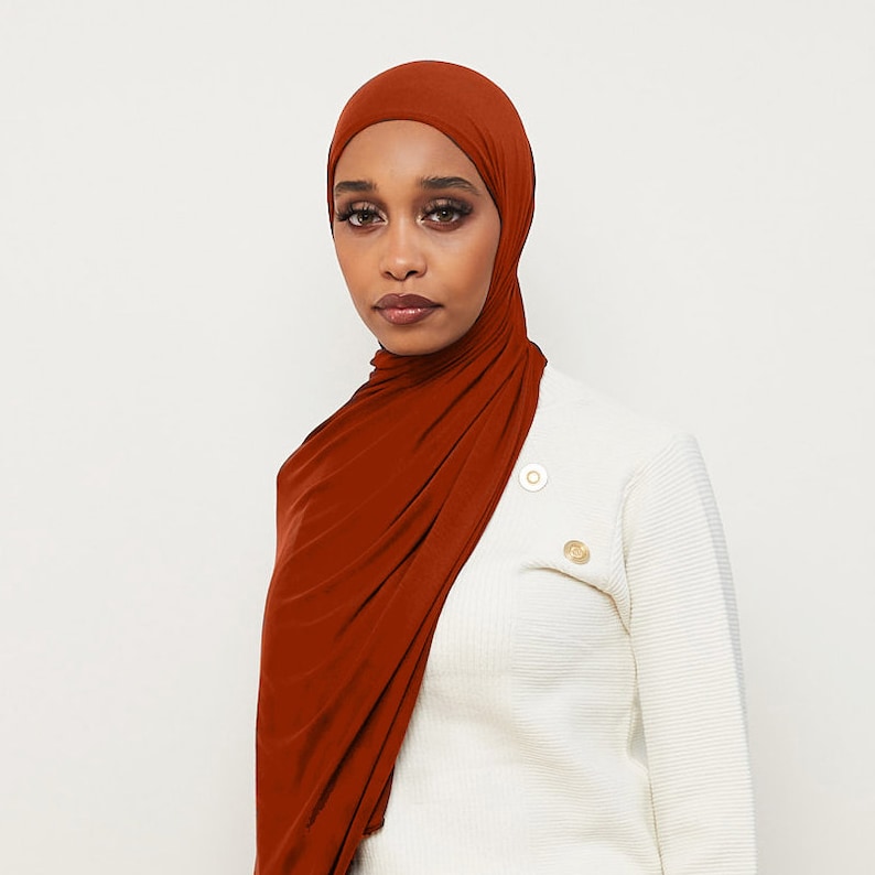 May include: A rust-colored jersey knit hijab with a simple, draped style. The hijab is worn by a person with dark skin and brown eyes. The person is wearing a white sweater.