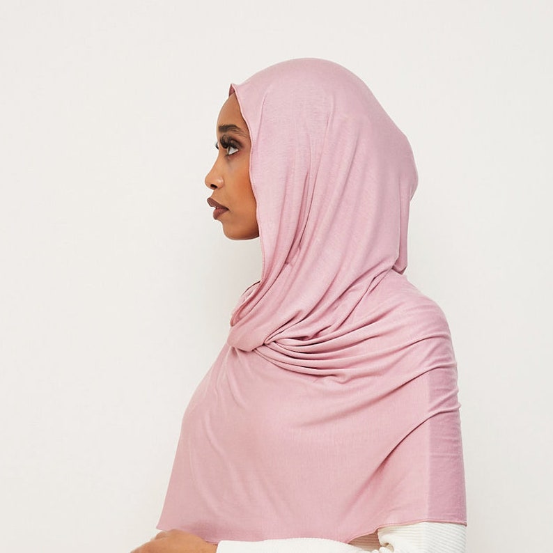 May include: A soft, pink hijab made of a stretchy fabric. The hijab is draped over the head and neck, with the ends tucked in at the shoulders.