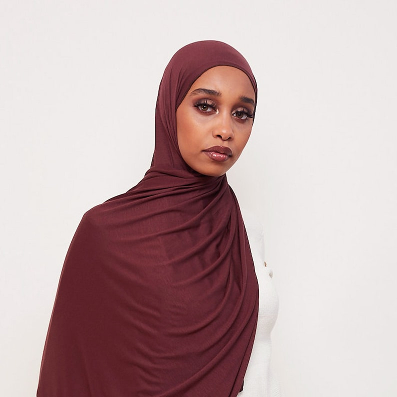 May include: A maroon-colored jersey knit hijab. The hijab is draped over the head and shoulders, with the fabric gathered in a loose, flowing style.