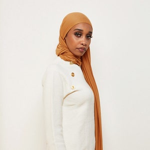 May include: A woman wearing a mustard yellow hijab with a white sweater. The sweater has gold buttons on the shoulder.