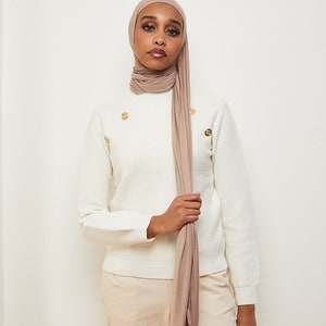 May include: A woman wearing a white sweater with gold buttons and a long, light brown scarf. She is wearing beige pants.