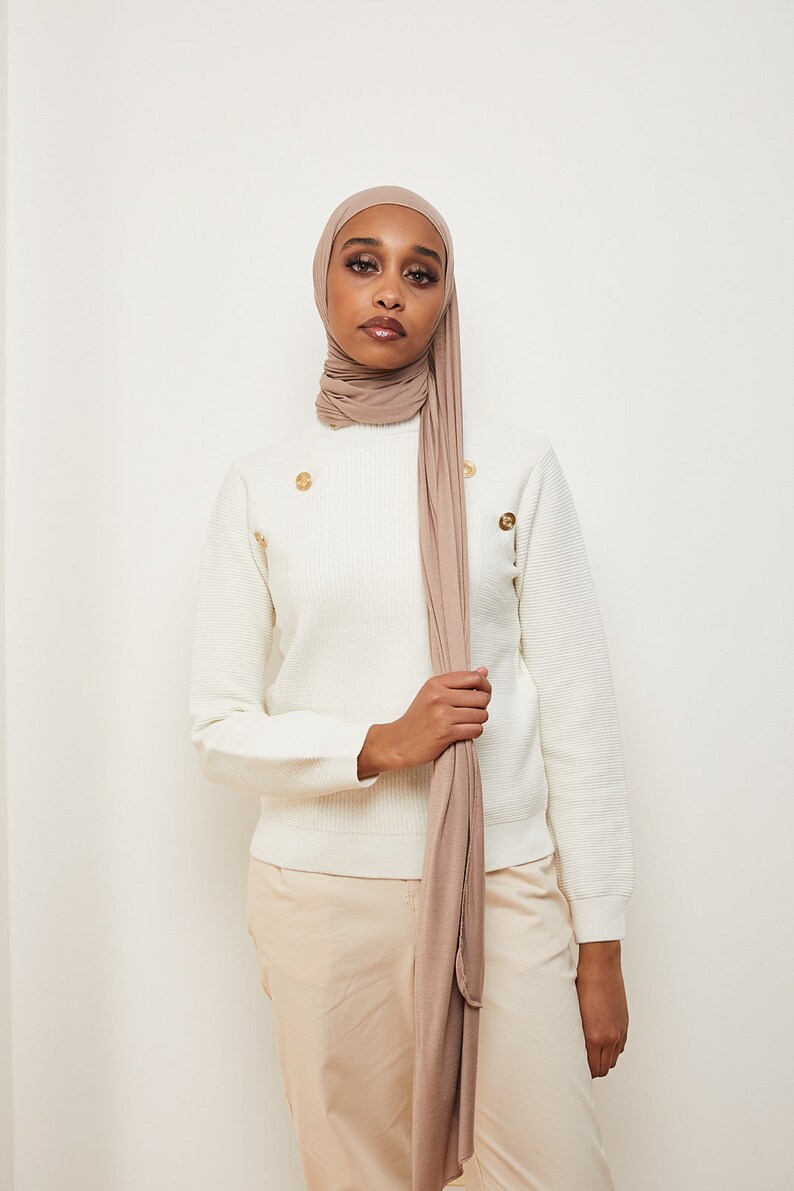 May include: A woman wearing a white sweater with gold buttons and a long, light brown scarf. She is wearing beige pants.
