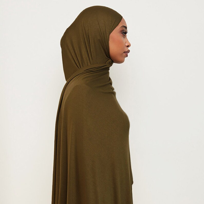 May include: A long, olive green hijab made of a soft, stretchy fabric. The hijab is draped over the head and shoulders, with the ends tucked in at the neck.