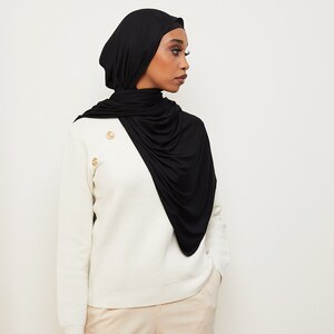 May include: A woman wearing a black jersey knit hijab with a white sweater and beige pants.