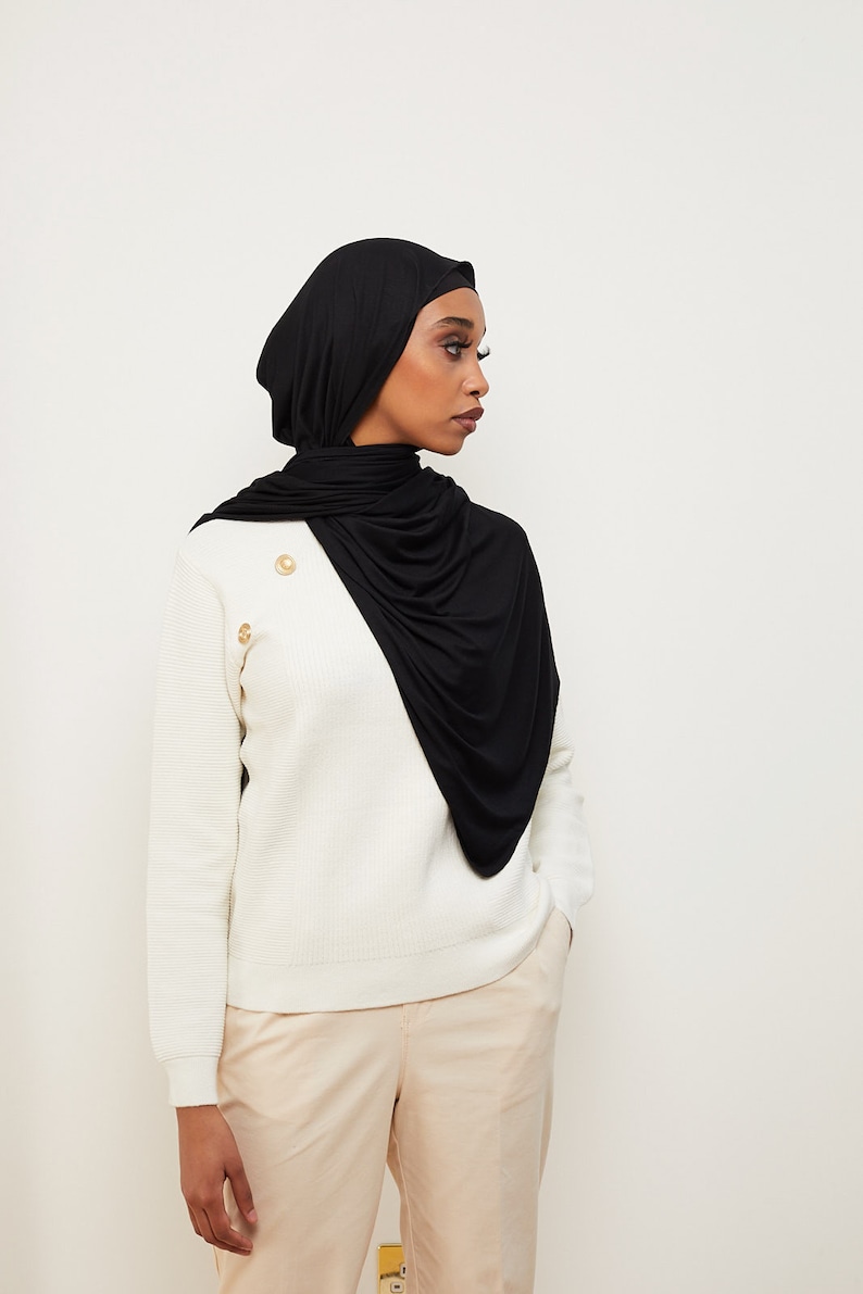 May include: A woman wearing a black jersey knit hijab with a white sweater and beige pants.