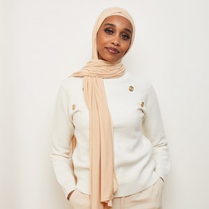 May include: A woman wearing a white sweater with gold buttons and beige pants. She is wearing a long, light beige scarf that is draped around her neck and shoulders.
