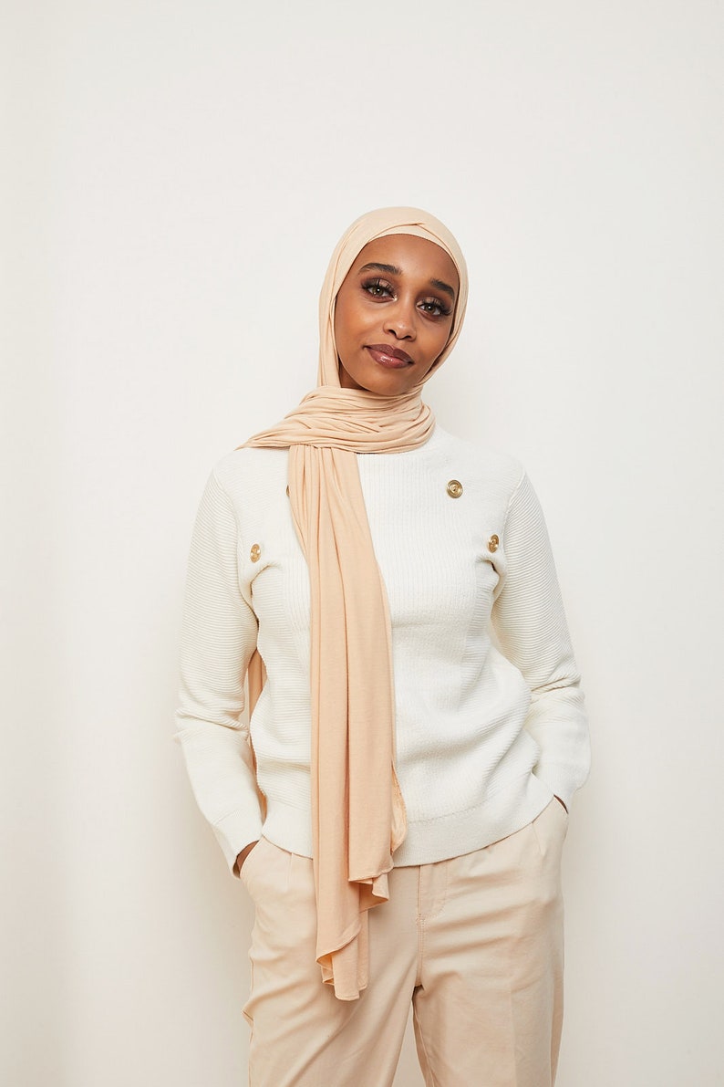 May include: A woman wearing a white sweater with gold buttons and beige pants. She is wearing a long, light beige scarf that is draped around her neck and shoulders.