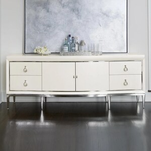 May include: A long, cream-colored sideboard featuring four drawers with silver-toned hardware and two central cabinet doors. The piece is supported by slender, tapered legs with a metallic finish and includes a rectangular tray holding glass bottles.