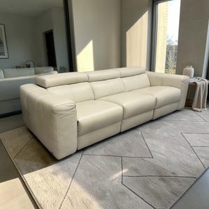 May include: A cream-colored leather sofa featuring three distinct seating sections, wide armrests, and adjustable headrests. The sofa has a smooth, uniform finish with visible stitching along the cushions and frame, providing a structured appearance.
