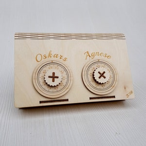 May include: A wooden box with two combination locks, one labeled "Oskars" and the other "Agnese". The box is made of light wood and has a natural finish. The locks are both round and have a black dial with white numbers. The box is dated "2018".
