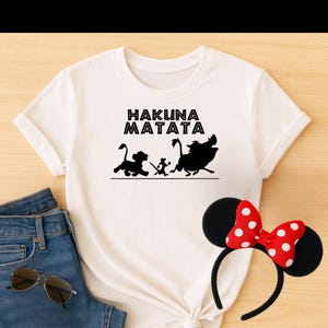 May include: White t-shirt with a black silhouette of Simba, Timon, and Pumbaa running. The text "Hakuna Matata" is printed above the silhouettes.