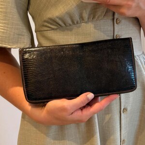 May include: A rectangular black wallet with a textured lizard-skin pattern and dark brown trim. The wallet is held horizontally, showcasing its slim, elongated shape and consistent dark finish across the front surface, highlighting the fine grain.