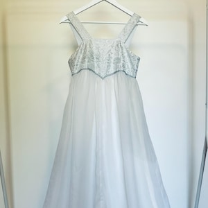Puede incluir: Un vestido formal blanco con corpiño bordado en plata y tirantes delicados cuelga de una percha blanca. El vestido presenta una falda larga y fluida. El vestido se exhibe en un perchero blanco sobre un fondo neutro.