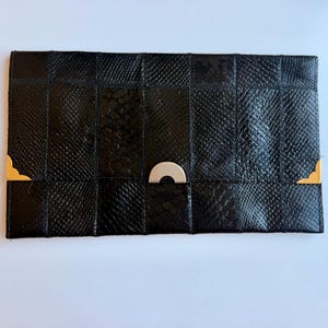 Puede incluir: Un bolso de mano rectangular negro con un diseño de patchwork que presenta diversas texturas de piel de serpiente. El accesorio está decorado con dos protectores de esquina de metal dorado y un cierre central de metal dorado semicircular.