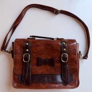 Può includere: Una borsa a tracolla marrone con un dettaglio a fiocco marrone scuro sulla patta anteriore, due cinghie verticali con fibbie e borchie color ottone, un manico superiore e una tracolla regolabile. La borsa ha una finitura liscia e lucida.