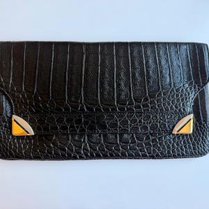 Può includere: Una pochette rettangolare nera con una trama testurizzata effetto pelle di coccodrillo. La patta anteriore è impreziosita da due angoli triangolari in metallo color argento posizionati sui lati inferiore sinistro e destro della chiusura.