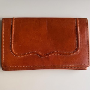 Puede incluir: Una cartera rectangular de cuero color coñac con un acabado liso y un panel decorativo cosido en forma de U en la solapa frontal. La cartera presenta costuras visibles del mismo tono a lo largo de los bordes y un diseño estructurado.
