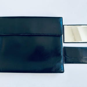 May include: A black leather clutch with a flap closure, a small matching wallet, and a rectangular mirror with a black frame. The items are arranged on a white surface.