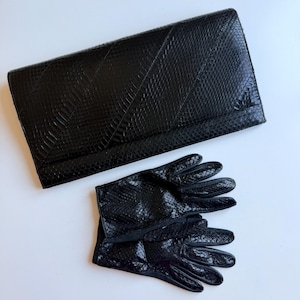 Könnte beinhalten: Eine schwarze, rechteckige Clutch mit einer strukturierten Schlangenhaut-Optik und ein passendes Paar schwarzer Schlangenhaut-Handschuhe. Die Clutch hat ein diagonales Nahtdesign auf der Vorderklappe, während die Handschuhe eine gleichmäßige Schuppenstruktur aufweisen.