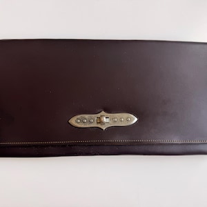Könnte beinhalten: Eine rechteckige, dunkelbraune Clutch mit glatter Oberfläche und sichtbaren Nähten an den Rändern. Die Mitte der Frontklappe ist mit einem länglichen, silberfarbenen Metallverschluss gesichert, der eine dekorative, strukturierte Oberfläche aufweist.