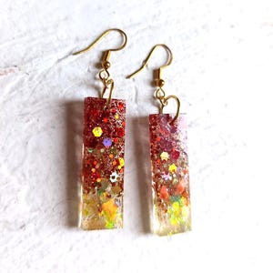 May include: A pair of rectangular earrings with gold-coloured hooks. The earrings are filled with red, gold, and multicoloured glitter, creating a sparkling effect. The earrings are a handmade jewellery item.