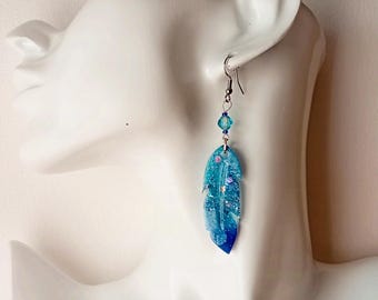 May include: A pair of dangle earrings featuring a blue and green feather-shaped charm with silver glitter accents. The earrings are made with a silver-toned metal and have a clear crystal bead.